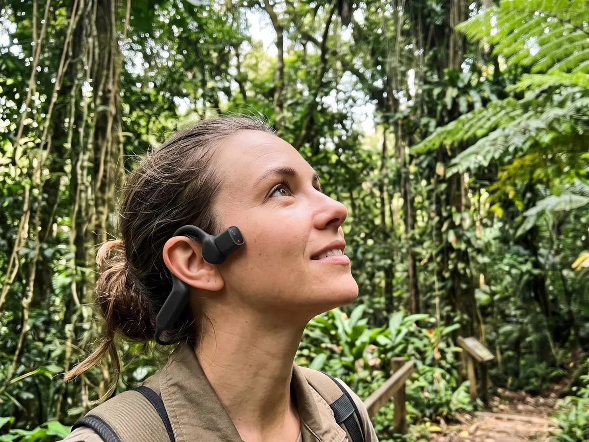A visitor wearing the black AiSee headset over one ear smiles up at the canopy on a rainforest trail.